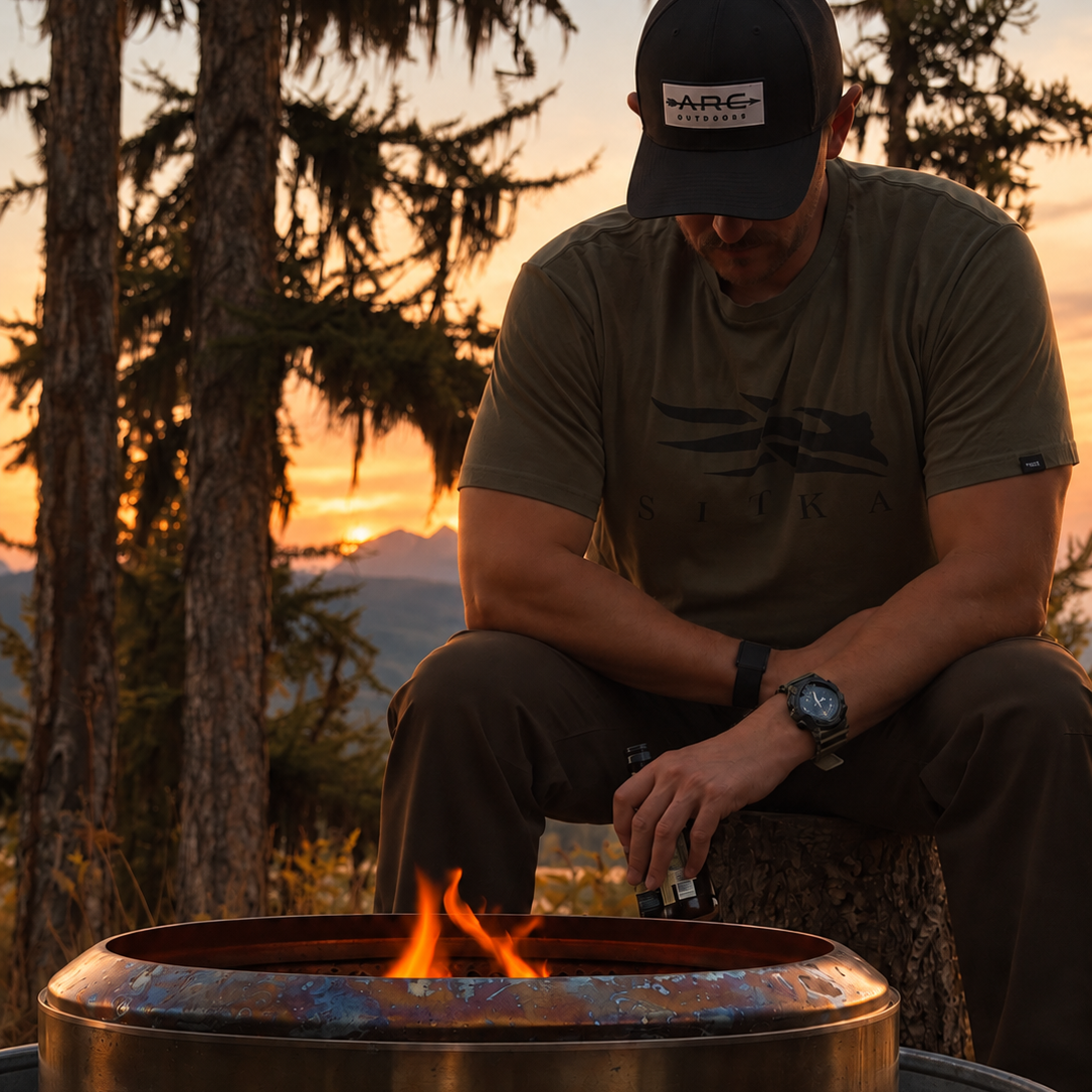 A man wearing a black ARC OUTDOORS cap and an olive green t-shirt sits by a metal fire pit with a small flame burning inside, set against a forest backdrop at sunset.