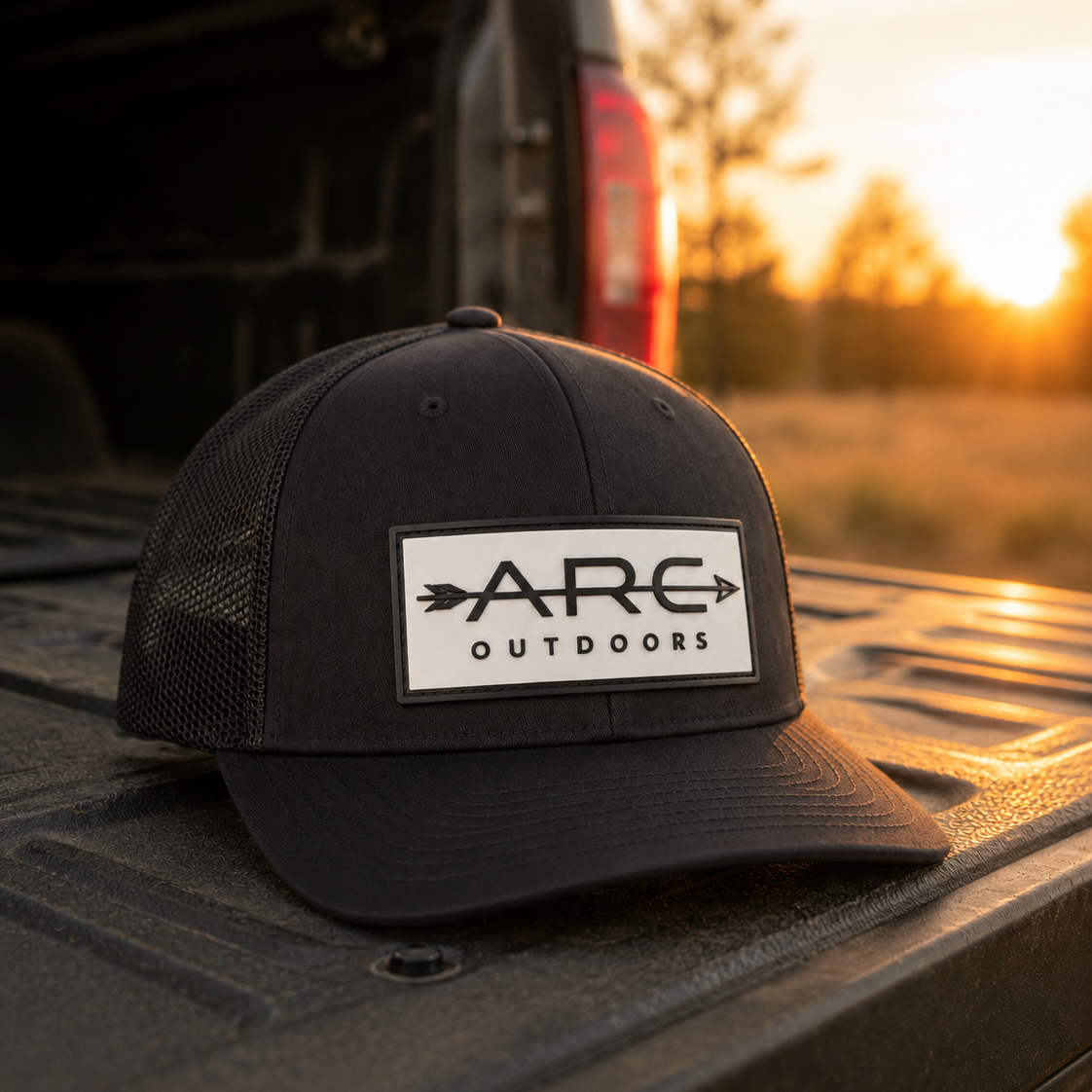 A black trucker-style cap with a white rectangular patch reading 'ARC OUTDOORS' sits on the tailgate of a pickup truck at sunset, with the truck bed and blurred outdoor scenery in the background.