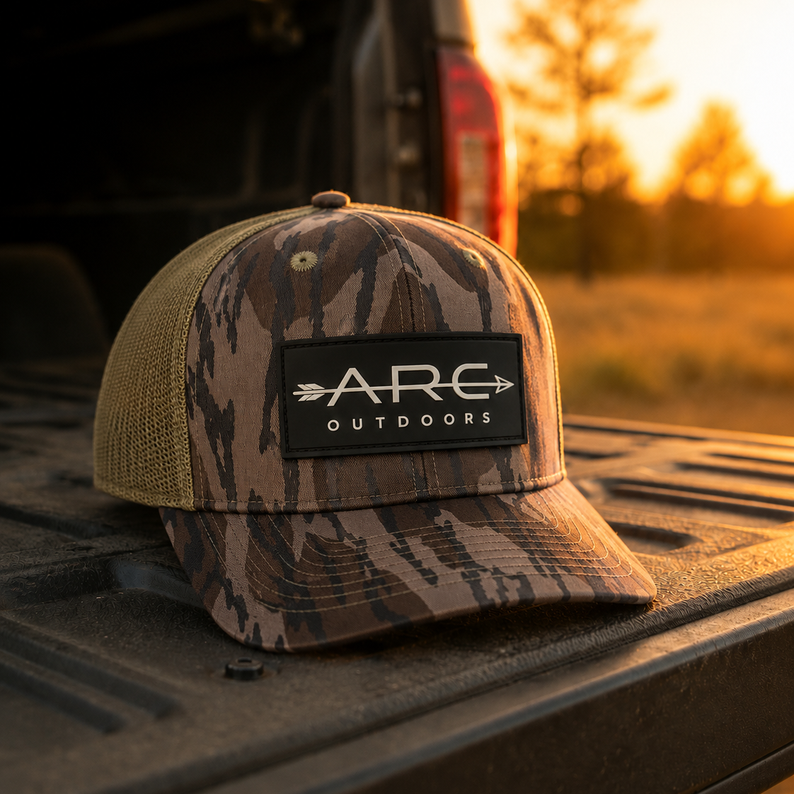 A camouflage trucker hat with a black patch reading 'ARC OUTDOORS' sits on the tailgate of a pickup truck at sunset, with a blurred outdoor field and trees in the background.